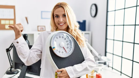 Beautiful blonde nutritionist woman holding weight machine to balance weight loss pointing thumb up to the side smiling happy with open mouth