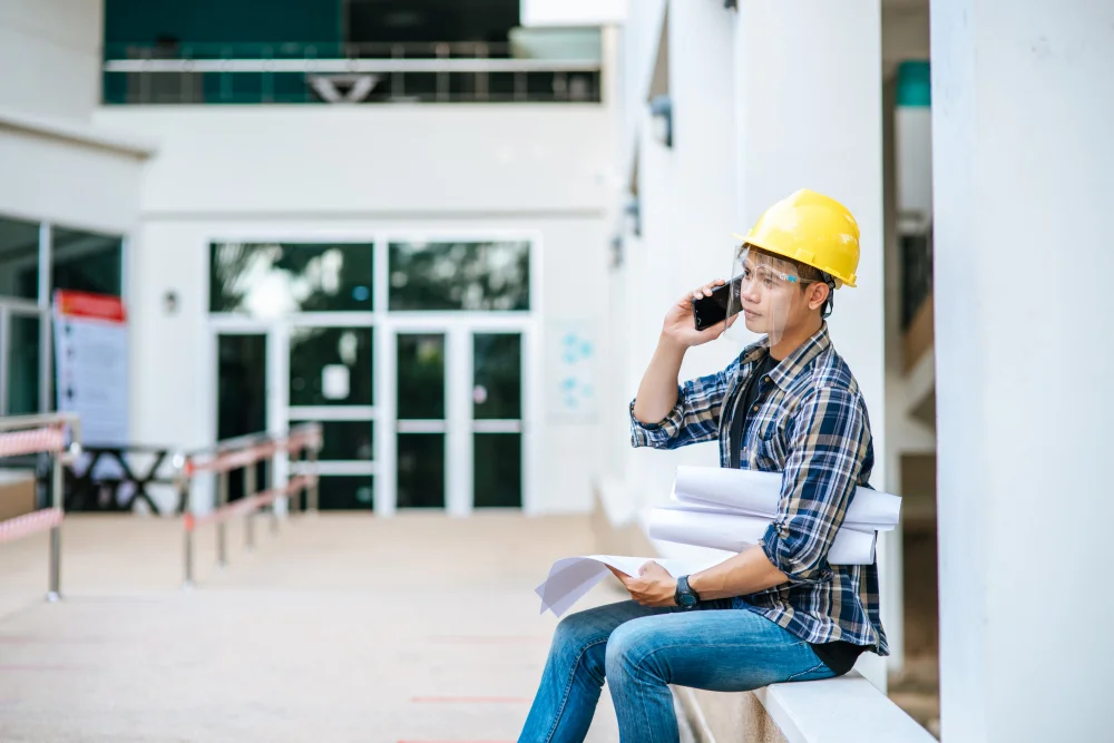 Construction worker wearing a yellow hard hat holding blueprints while talking on the phone at a building site.