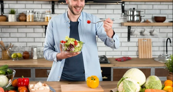 Happy man standing in the kitchen eating fresh salad with fork-eat this and live