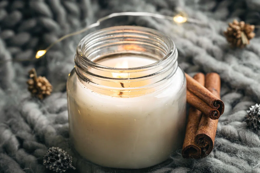 Lit candle in glass jar on knit blanket with cinnamon, pinecones, and string lights.