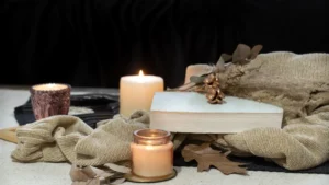 Lit candles with a book, dried leaves, and beige blanket create a warm autumn atmosphere.
