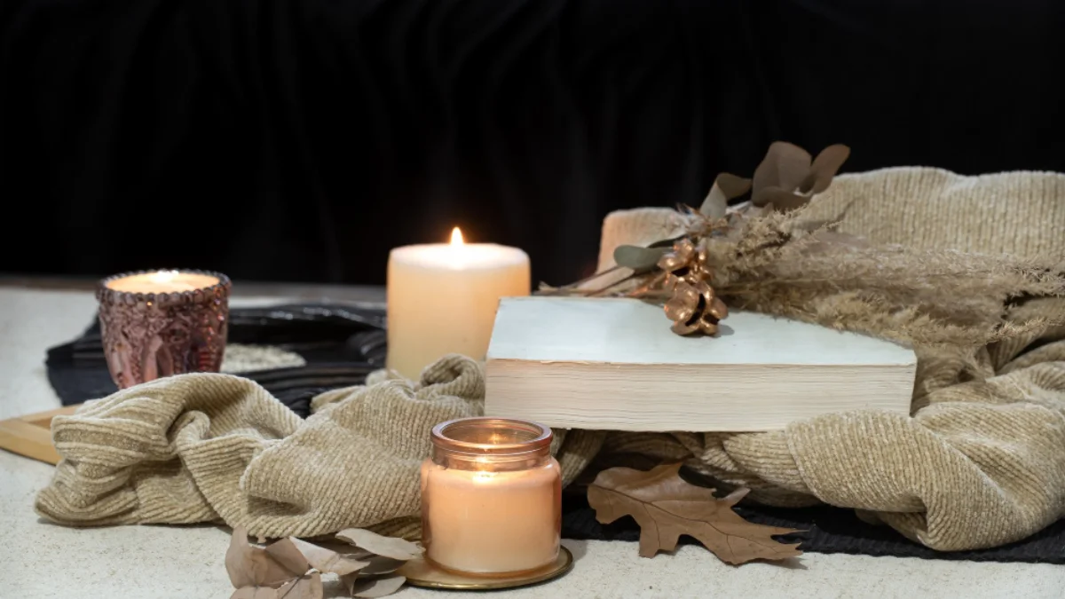 Lit candles with a book, dried leaves, and beige blanket create a warm autumn atmosphere.