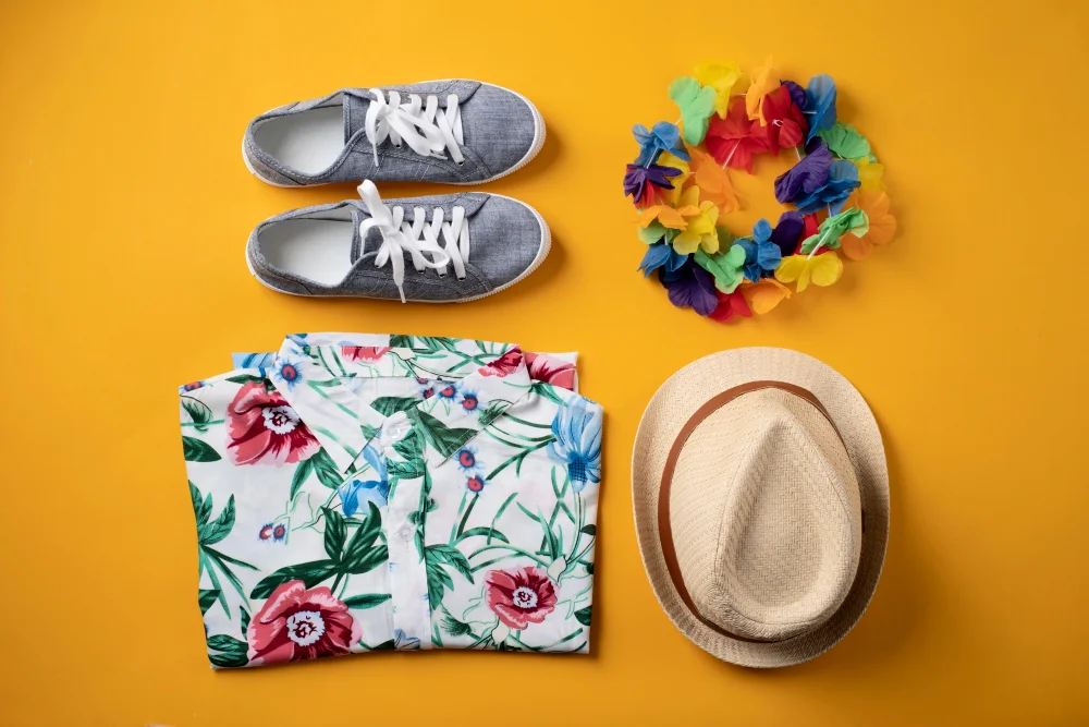Summer outfit with sneakers, Hawaiian shirt, hat, and colorful lei on yellow background.