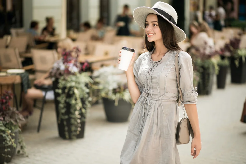 Woman in gray dress and hat holding coffee outside a café.